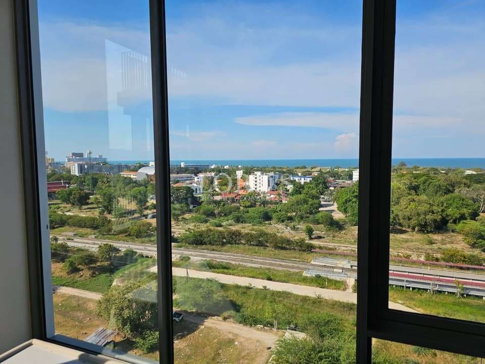 High-floor apartment window view overlooking green landscape, railway tracks, city buildings, and the ocean under a clear blue sky.