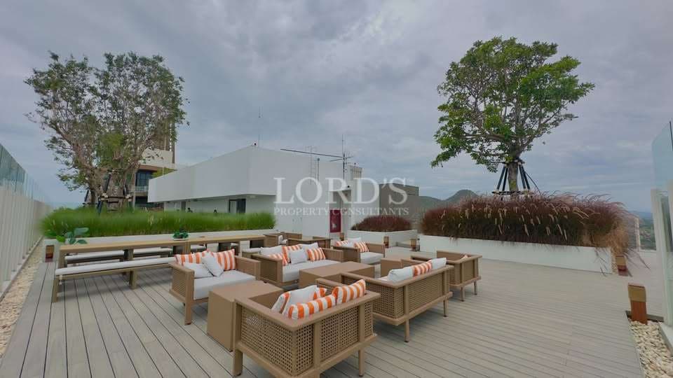 Luxury rooftop lounge with modern outdoor seating, wooden deck flooring, landscaped greenery, glass railings, and panoramic city views under a cloudy sky.