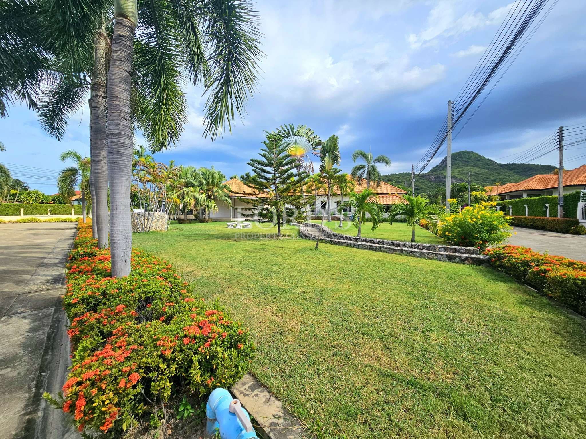 Well-maintained residential garden with palm trees, green lawn, flower beds, and mountain backdrop