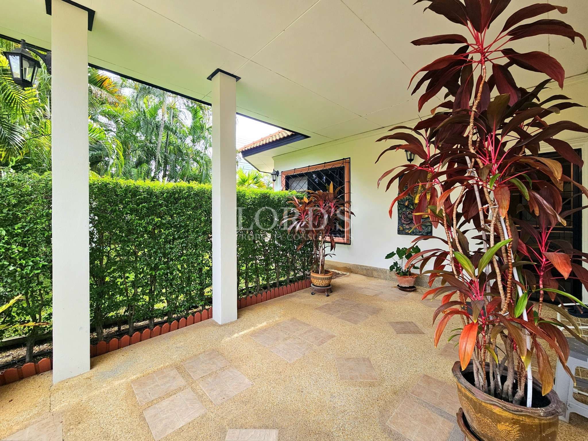 Covered veranda with potted plants, tiled flooring, white pillars, and lush garden hedge
