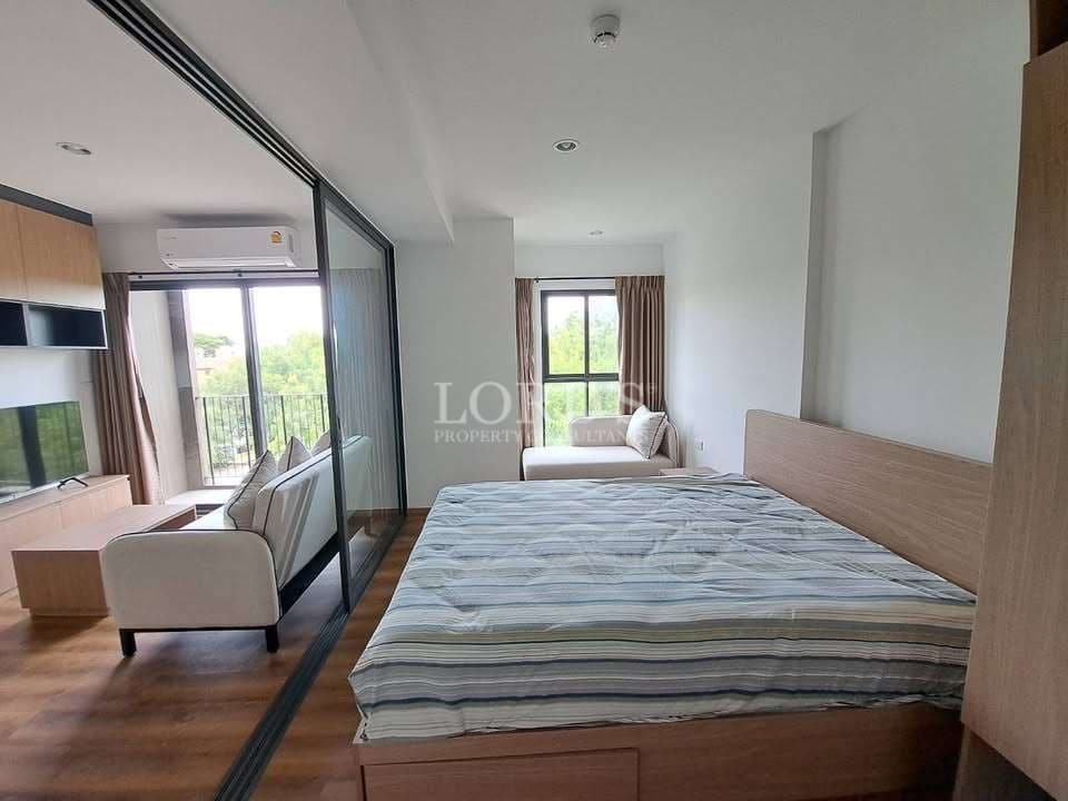 Modern one-bedroom condo with glass sliding partition, wooden bed frame, cozy seating area, and large window with greenery view.