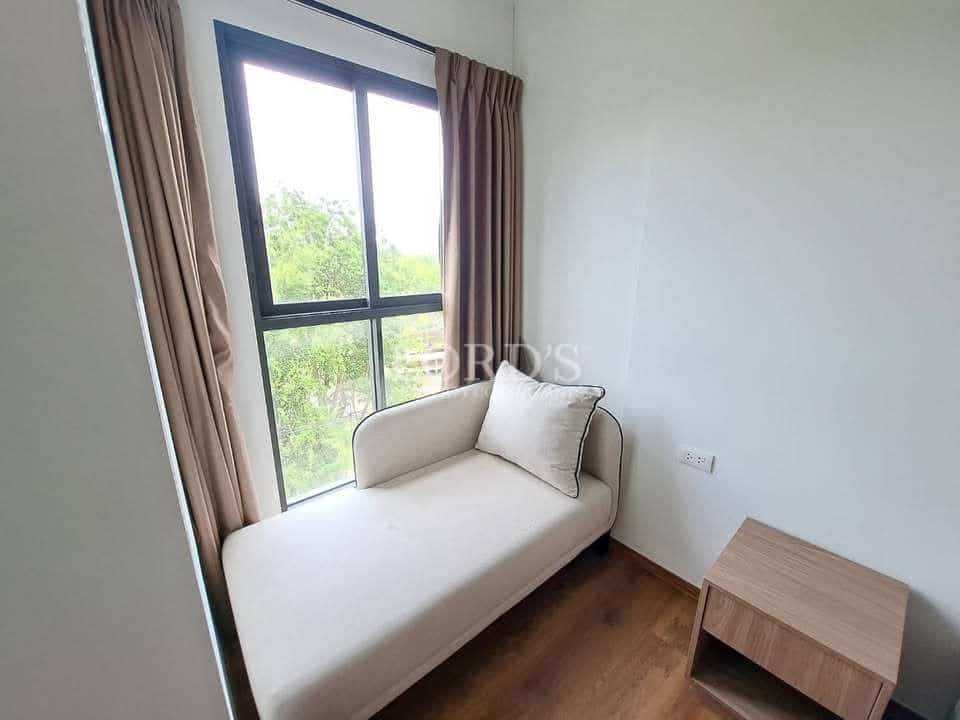 Minimalist bedroom corner with single daybed, cushion, wooden side table, and large window with curtains.