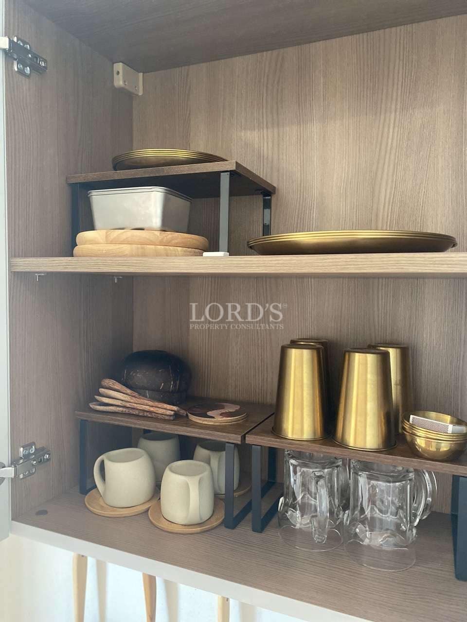 Organized kitchen cabinet with wooden shelves, cups, glasses, plates, and gold-toned kitchenware.