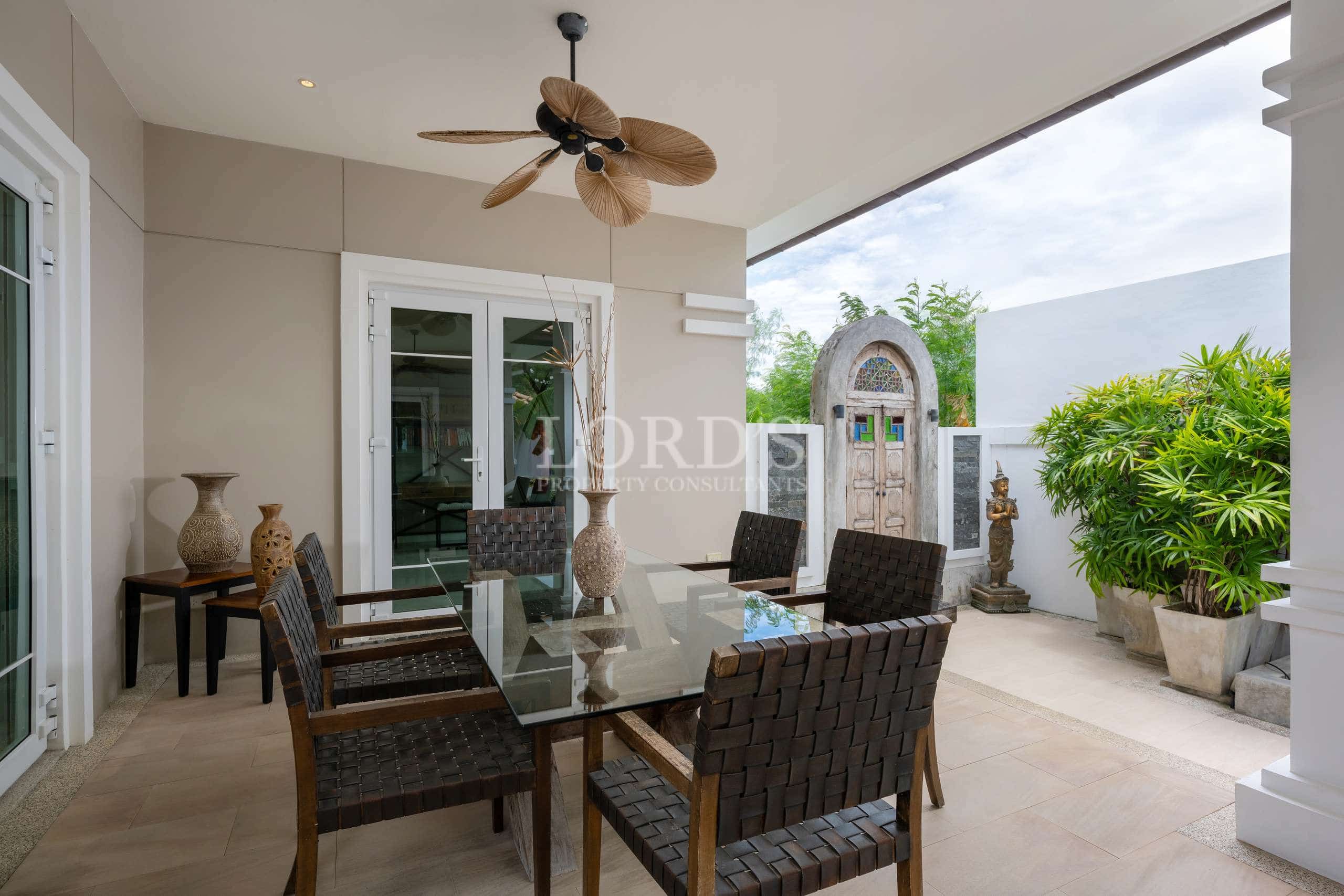 Covered outdoor dining patio with glass table, wicker chairs, ceiling fan, and garden view in a luxury villa.