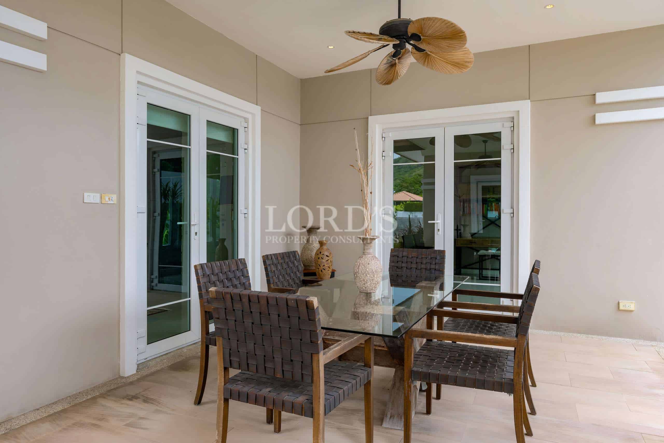 Covered outdoor dining patio with glass table, woven chairs, ceiling fan, and French doors in a luxury villa.