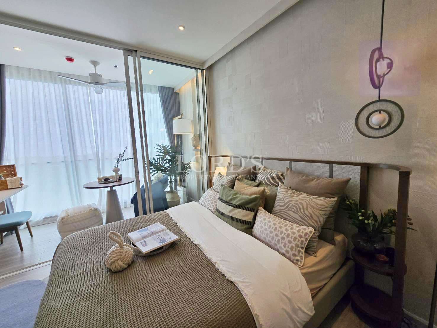 Modern bedroom with cozy bed, decorative pillows, pendant lighting, textured wall, and glass sliding doors opening to a balcony seating area.