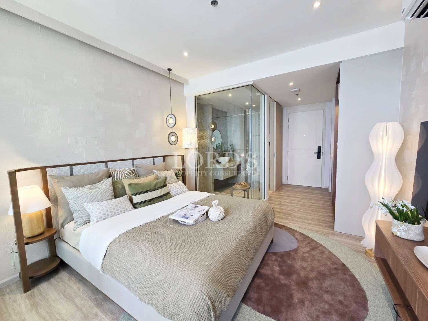 Modern bedroom interior with neutral decor, double bed, decorative pillows, bedside lighting, glass-enclosed bathroom, wooden flooring, and minimalist furniture.