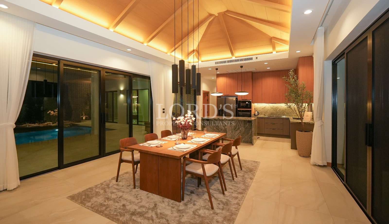 Luxury open-plan dining and kitchen area with wooden ceiling, pendant lighting, and floor-to-ceiling glass doors.