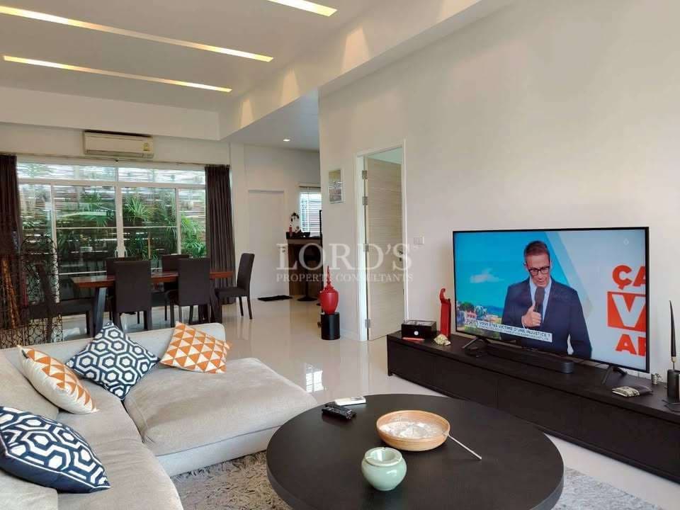 Modern living room with sofa seating, large TV, coffee table, recessed ceiling lights, and open dining area.