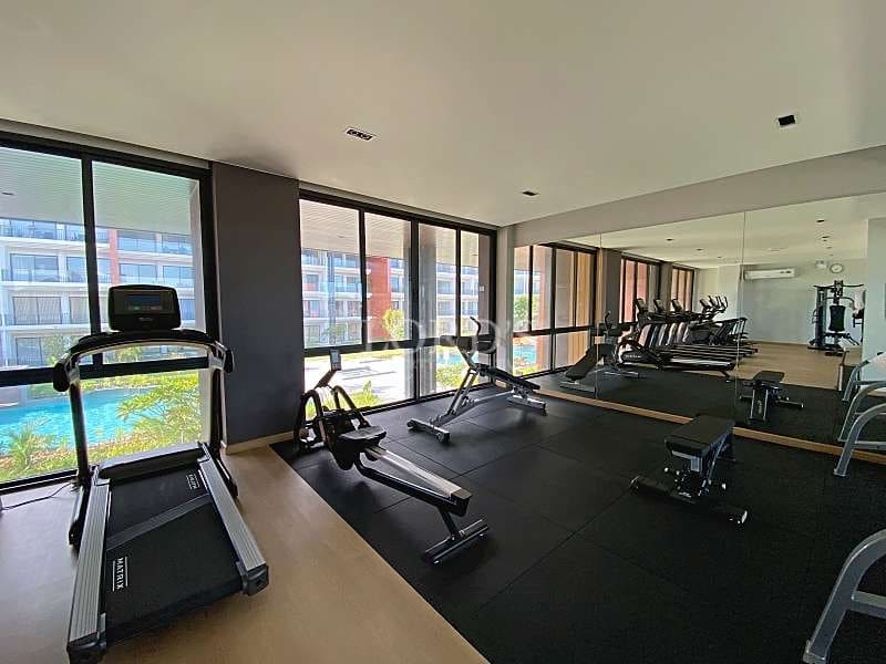 Modern apartment gym with treadmills, rowing machine, weight benches, mirrored wall, and large windows overlooking the pool.