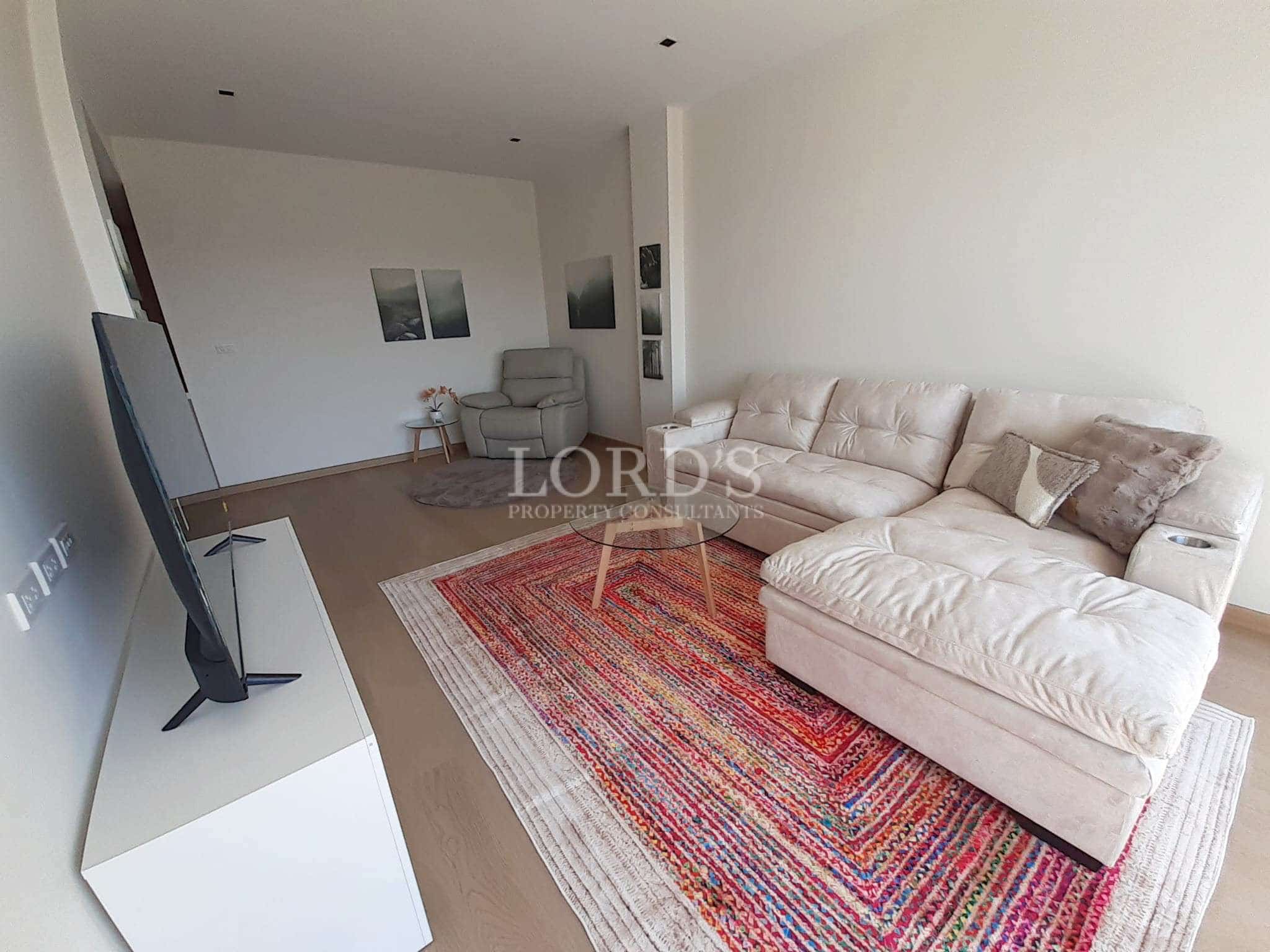 Modern living room with sectional sofa, wall-mounted TV, wooden floor, colorful area rug, and minimalist décor