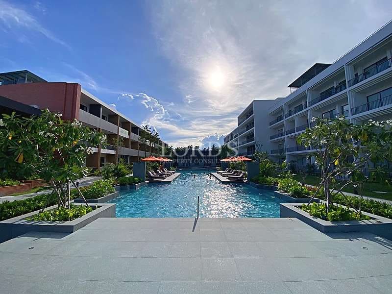 Resort-style swimming pool between modern apartment buildings with sun loungers, umbrellas, and landscaped greenery.