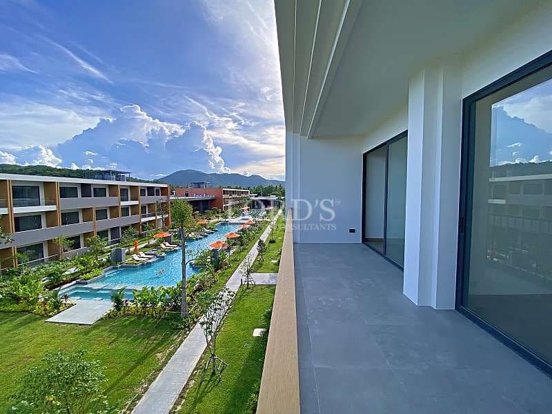 Apartment balcony overlooking a resort-style swimming pool, landscaped gardens, modern buildings, and mountain views.
