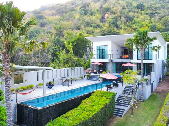 Luxury modern villa with an infinity swimming pool, outdoor terraces, and palm trees set against a lush hillside backdrop.