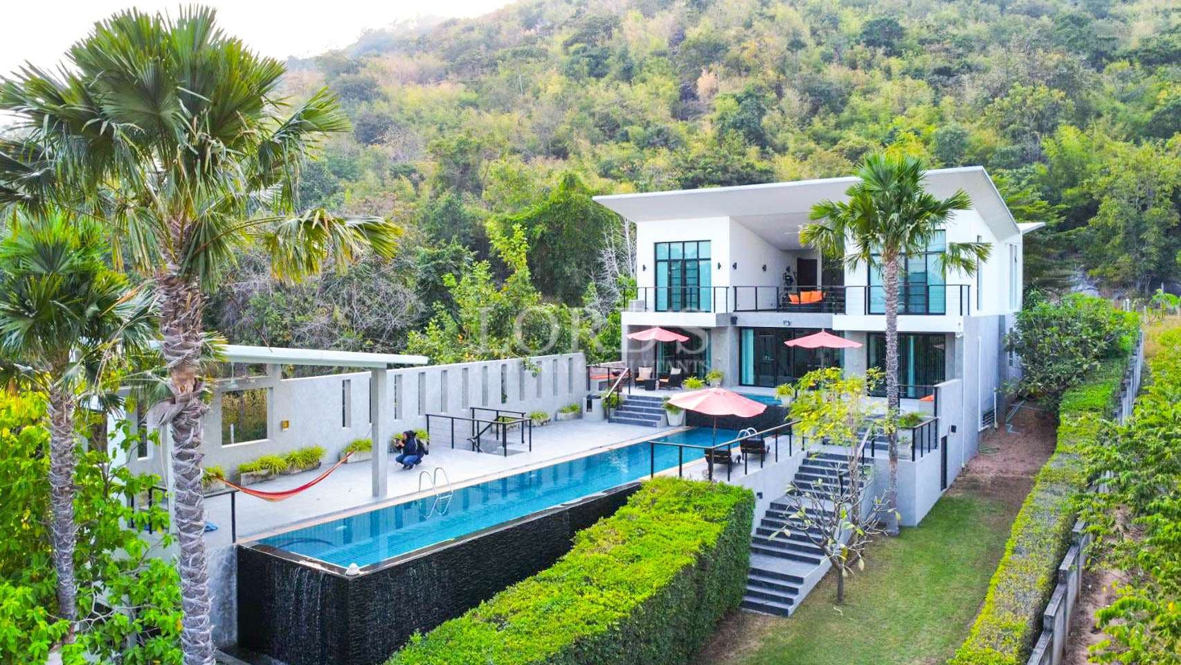 Luxury modern villa with an infinity swimming pool, outdoor terraces, and palm trees set against a lush hillside backdrop.