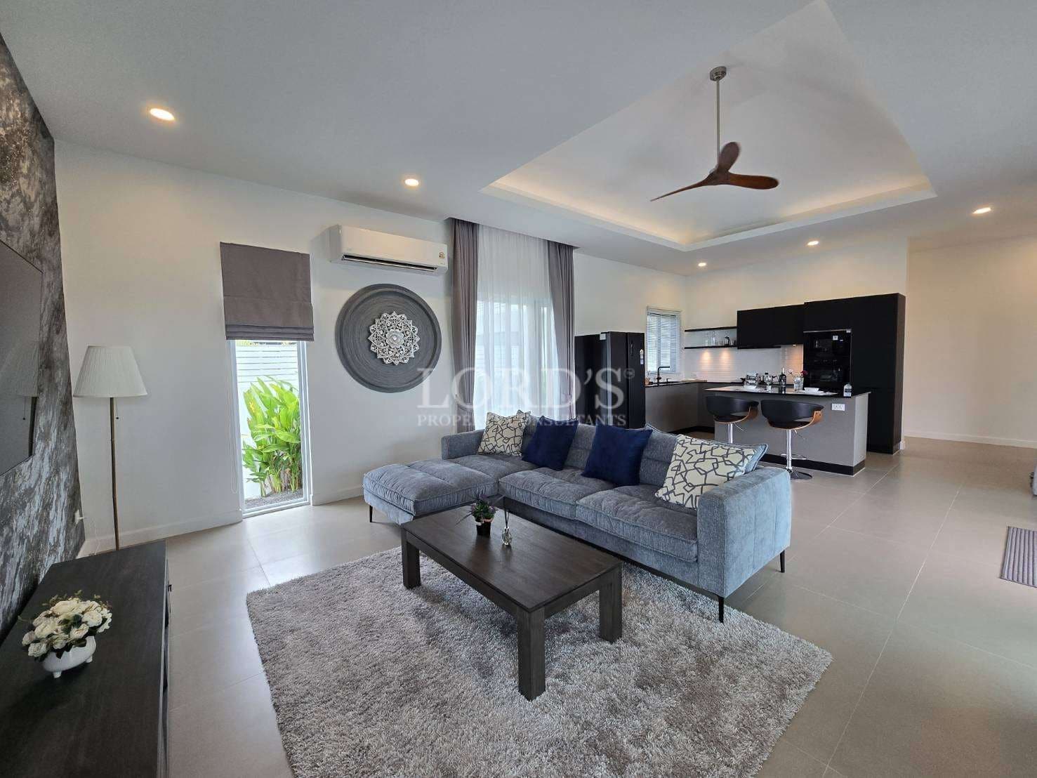 Open-plan modern living room with sectional sofa, ceiling fan, and contemporary kitchen.