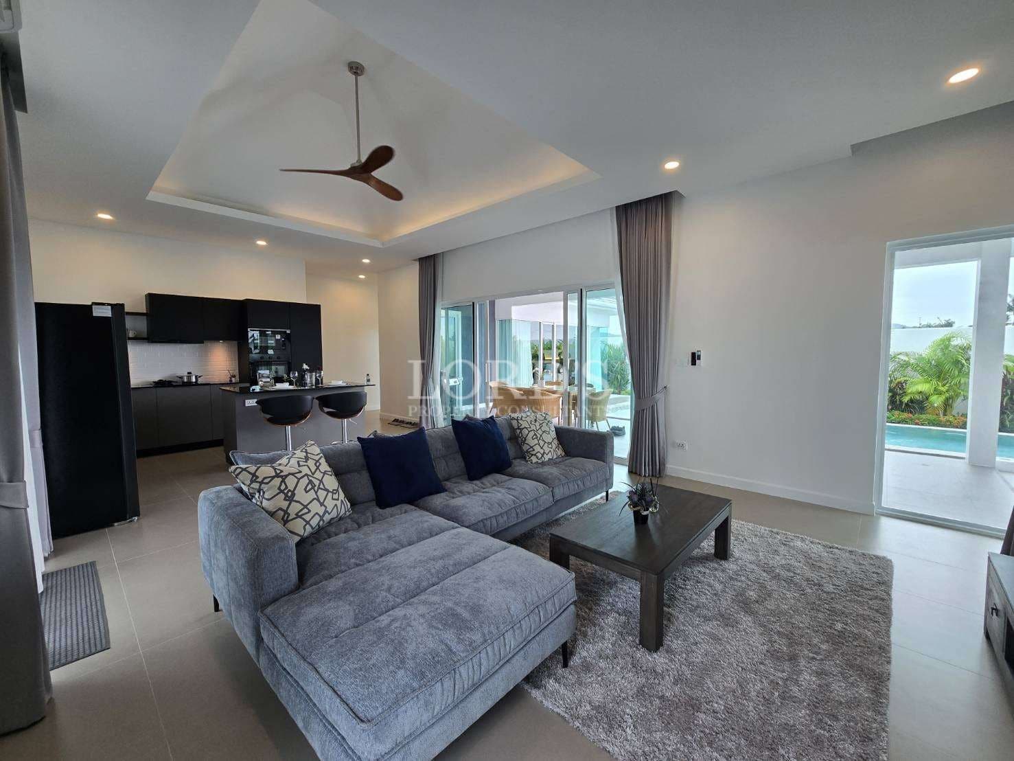 Modern open-plan living room with sectional sofa, ceiling fan, and kitchen view.