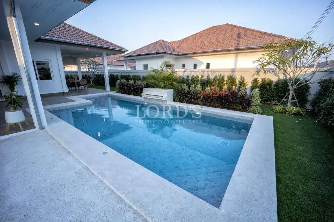 Modern private swimming pool with white stone decking and landscaped garden beside a covered villa patio.