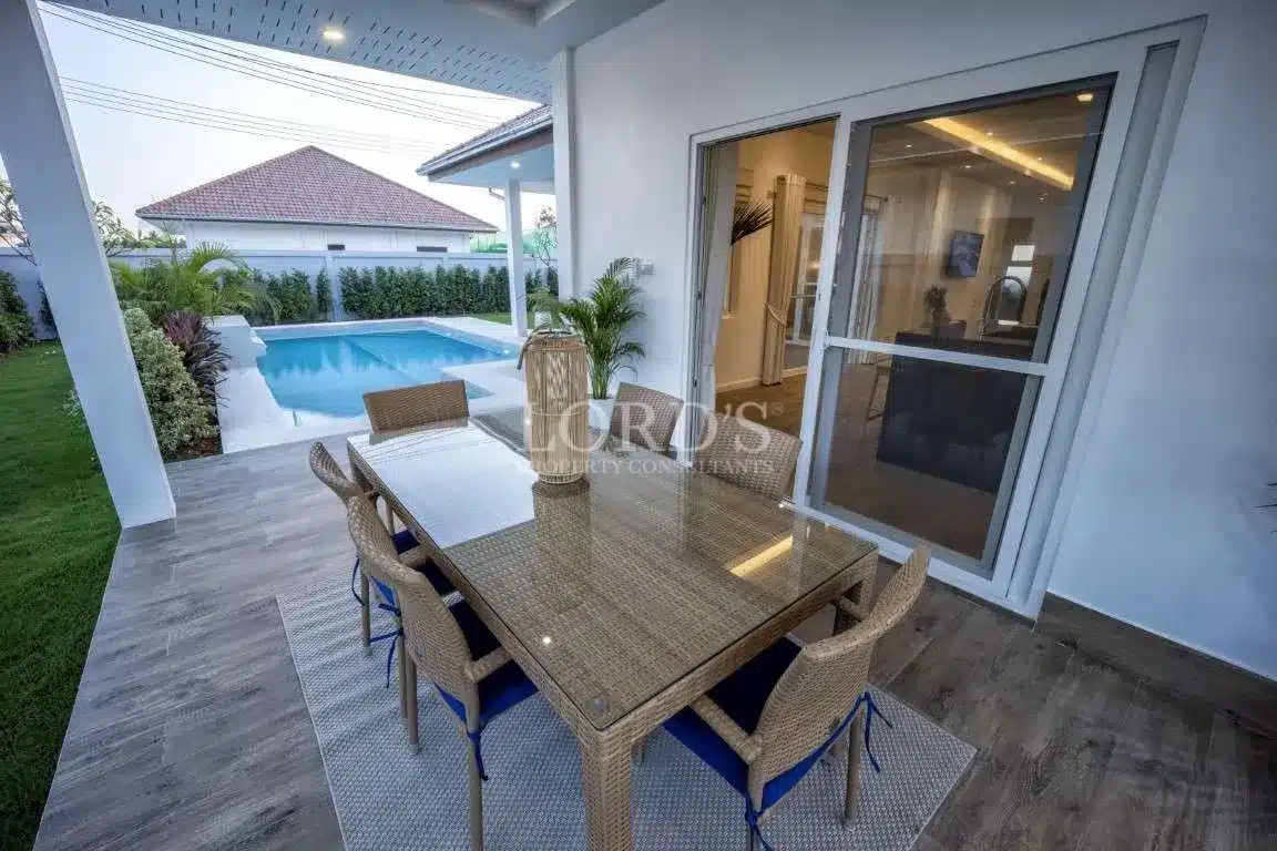 Covered outdoor dining patio with wicker table and chairs overlooking a private swimming pool and garden, connected to the home by sliding glass doors.