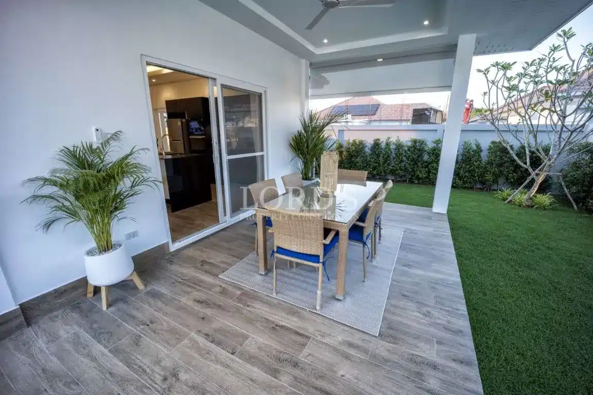 Covered outdoor patio with wooden flooring, glass dining table, wicker chairs, sliding glass doors, and a landscaped garden area.