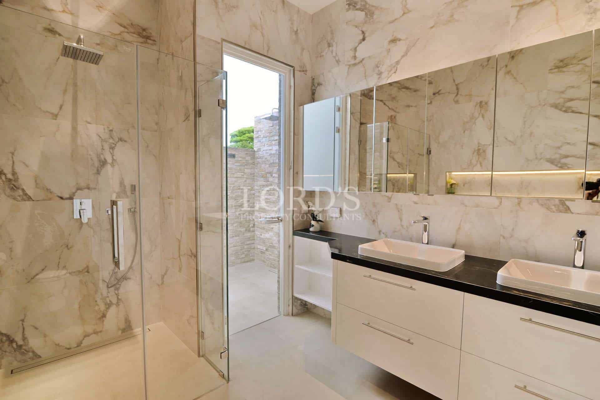 Modern luxury bathroom with marble walls, glass rain shower, double vanity, and access to a private outdoor shower.