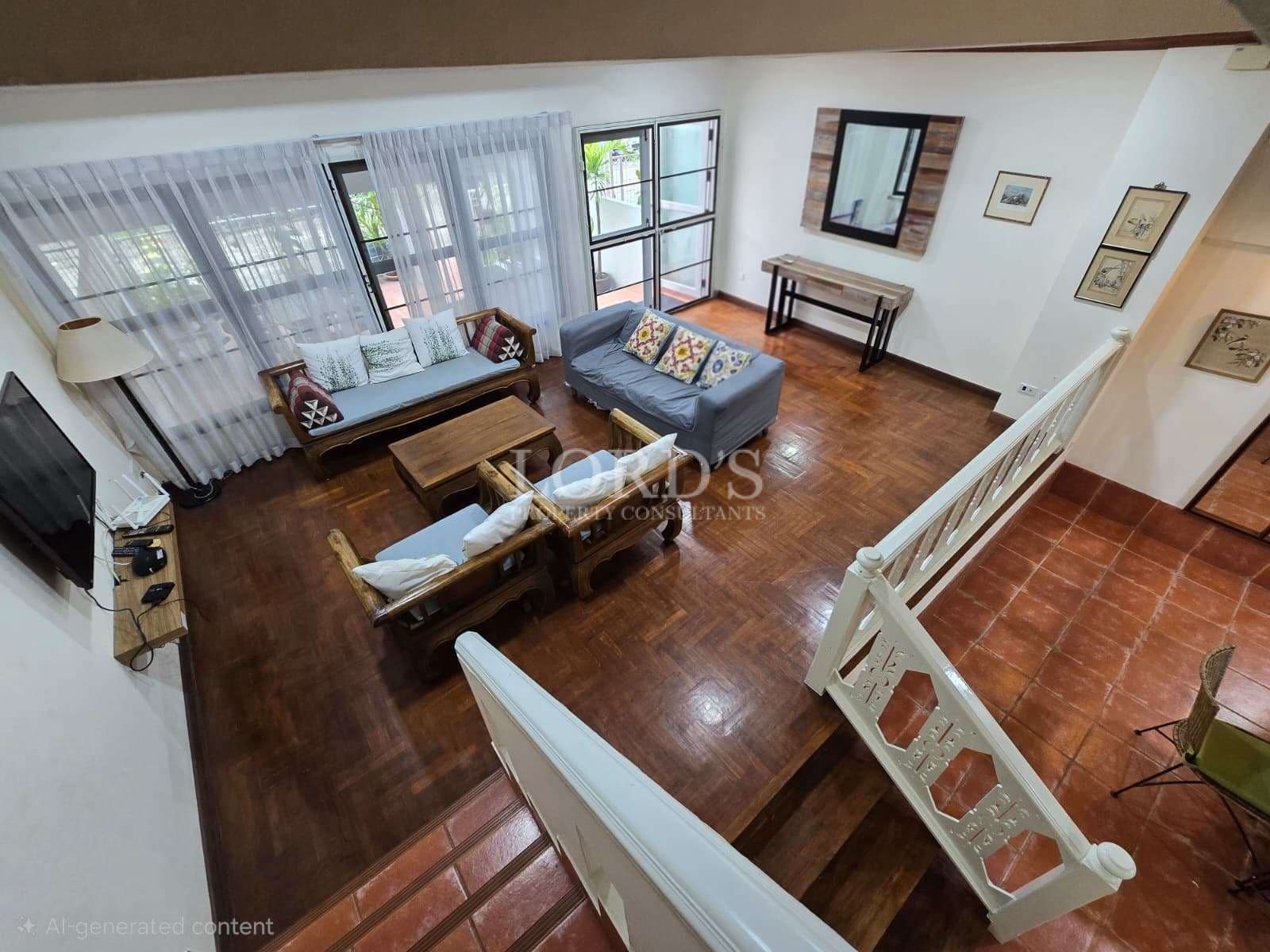 Open-plan living area with wooden flooring, seating arrangement, staircase railing, and large windows.