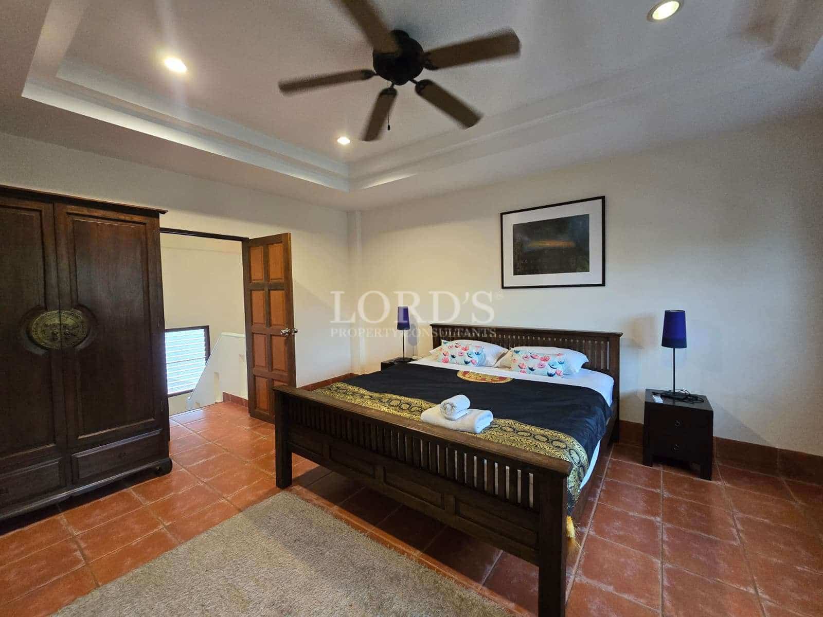 Spacious bedroom with wooden bed, ceiling fan, wardrobe, terracotta tile floor, and bedside lamps.
