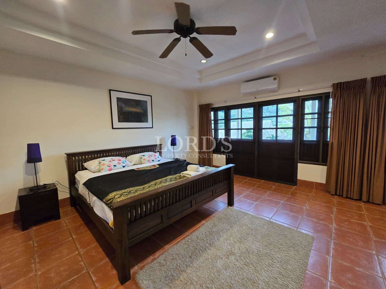 Spacious bedroom with wooden bed, ceiling fan, terracotta tile flooring, and large window doors.