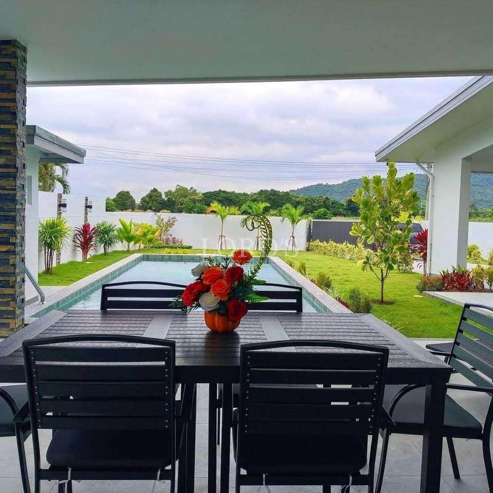 Outdoor patio dining area overlooking a private swimming pool, landscaped garden, and modern villa with green lawn and scenic hill views.