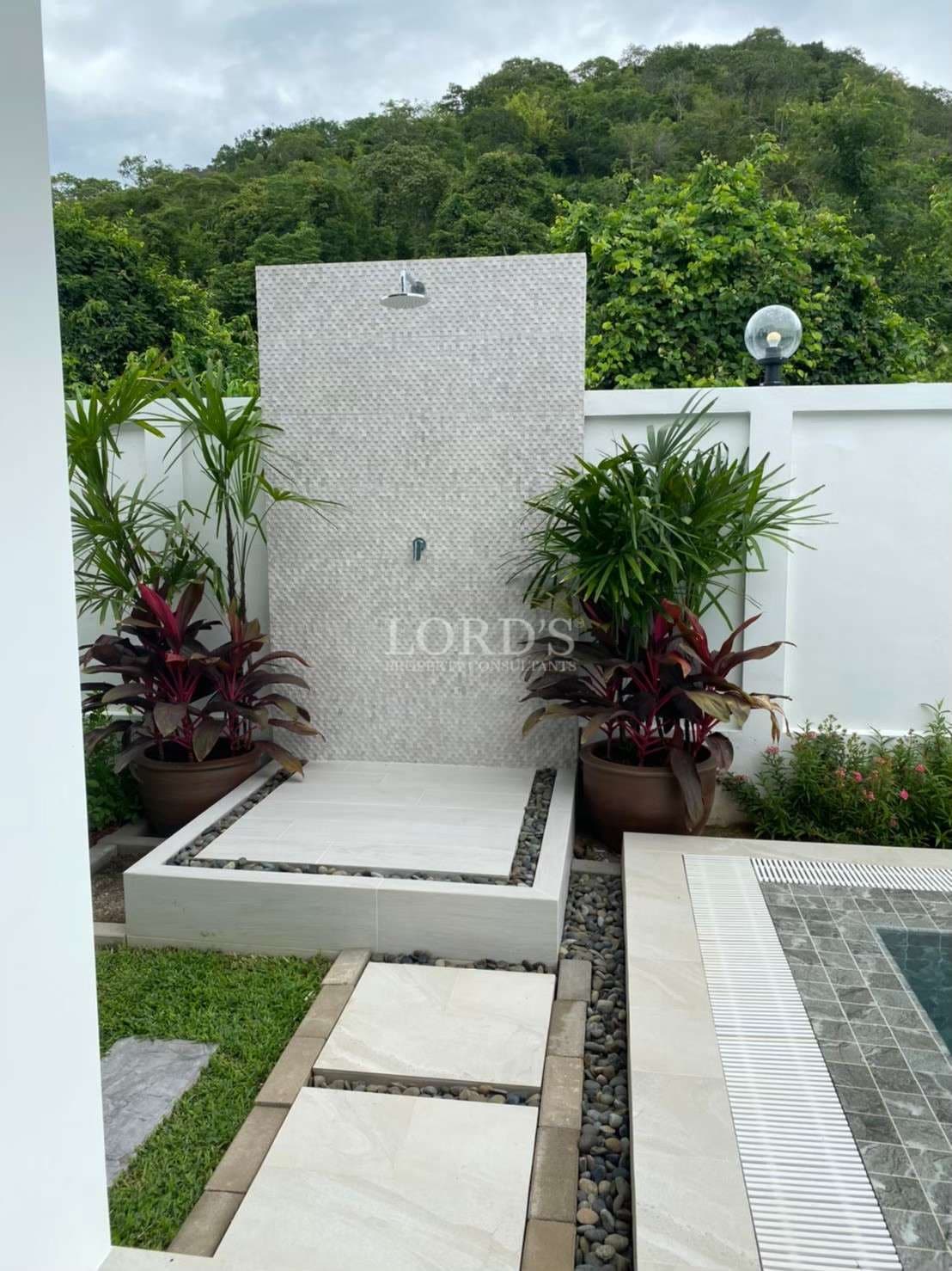 Outdoor garden shower beside a private swimming pool with stone-tiled wall, tropical plants, and hillside backdrop.