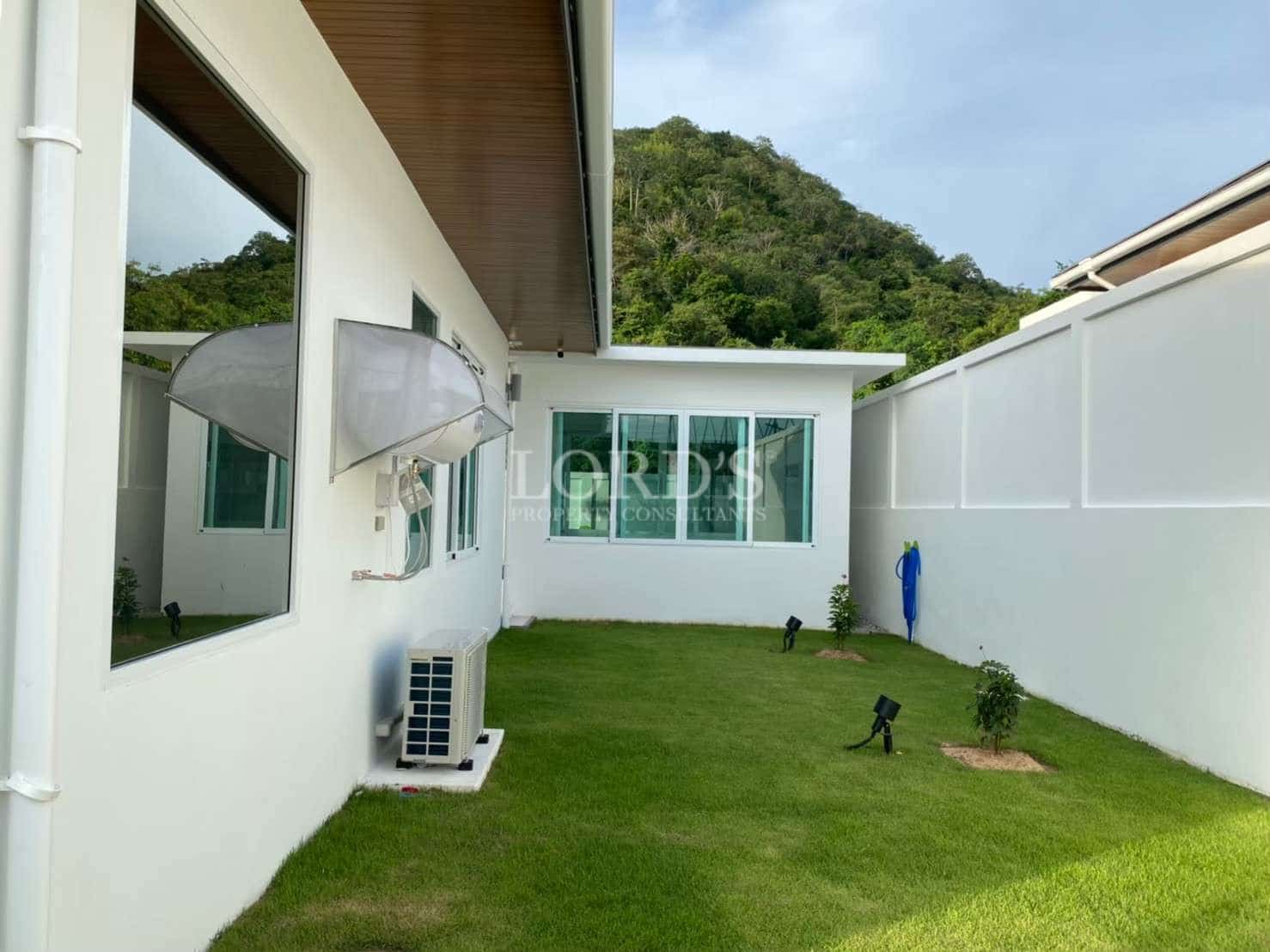 Side yard of a modern villa with green lawn, white perimeter walls, exterior lighting, and air conditioning units.