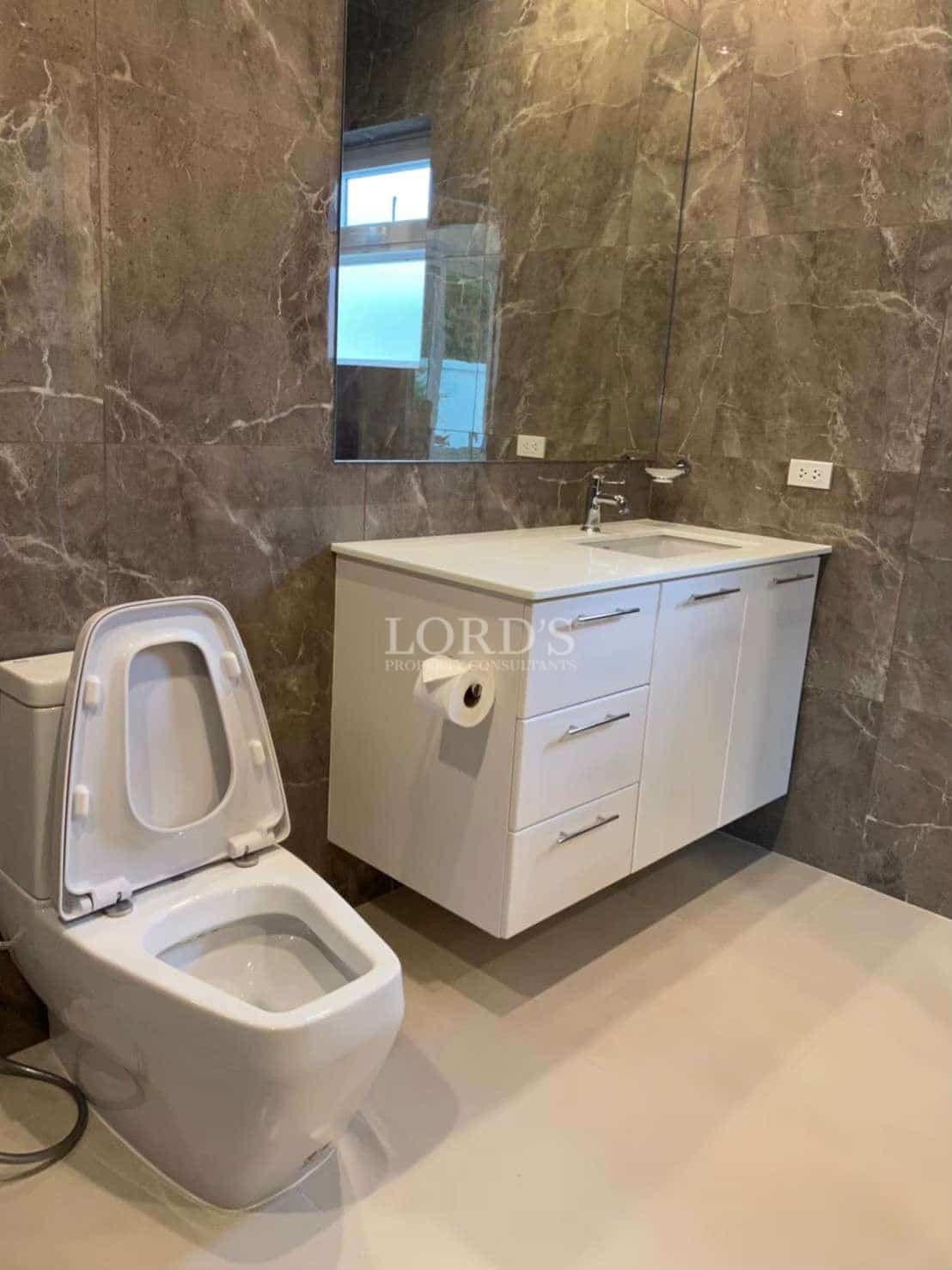 Modern bathroom with white vanity cabinet, wall mirror, marble-look tiled walls, and contemporary toilet.