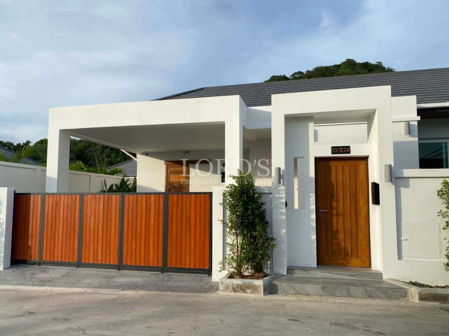 Modern villa exterior with a white minimalist facade, wooden gate, covered carport, and private entrance.