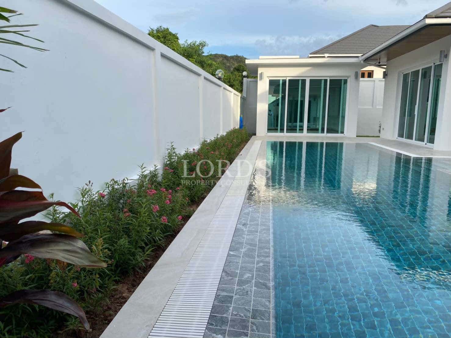 Private swimming pool beside a modern villa with blue tiled water, landscaped garden, and white perimeter wall.