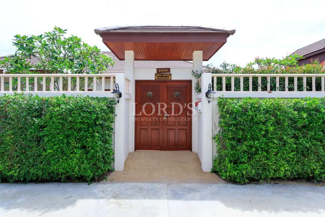 Private villa entrance with a wooden double gate, white perimeter wall, manicured green hedges, and a covered entryway.