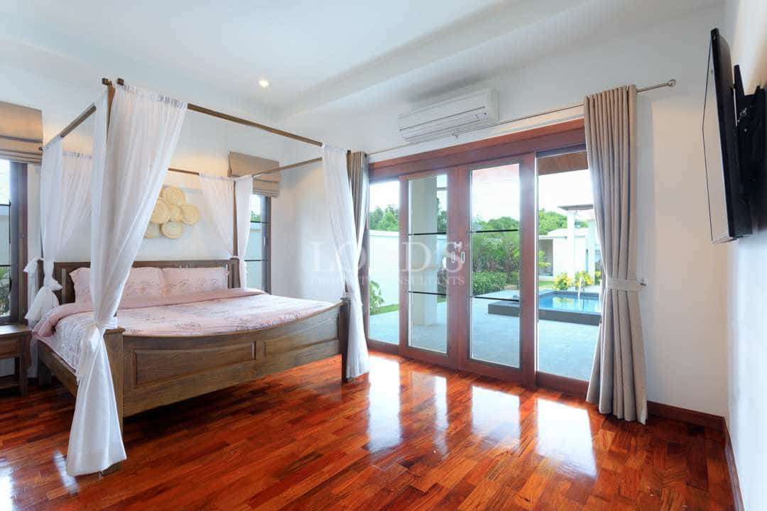 Spacious luxury bedroom with a wooden four-poster bed, sheer canopy curtains, polished wooden floors, and large glass doors opening to a pool area.