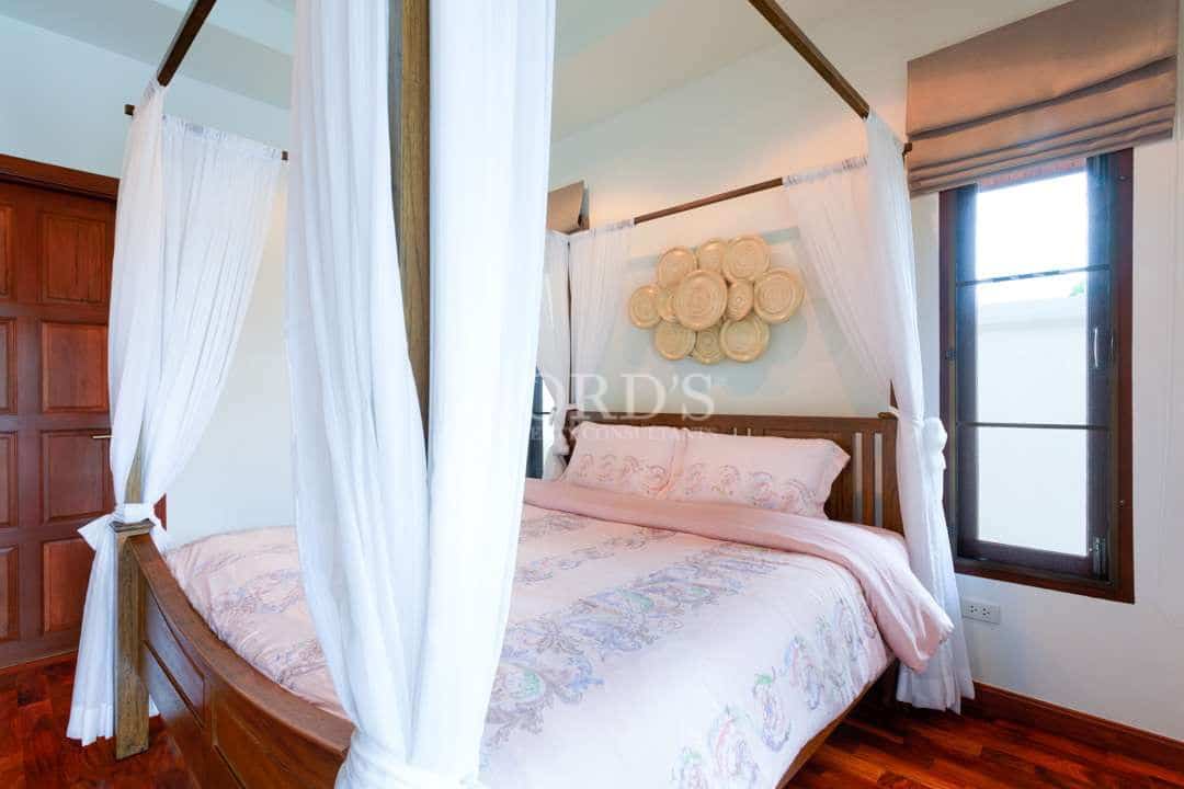 Elegant bedroom with a wooden four-poster bed, sheer white canopy curtains, soft pastel bedding, and natural light from tall windows.