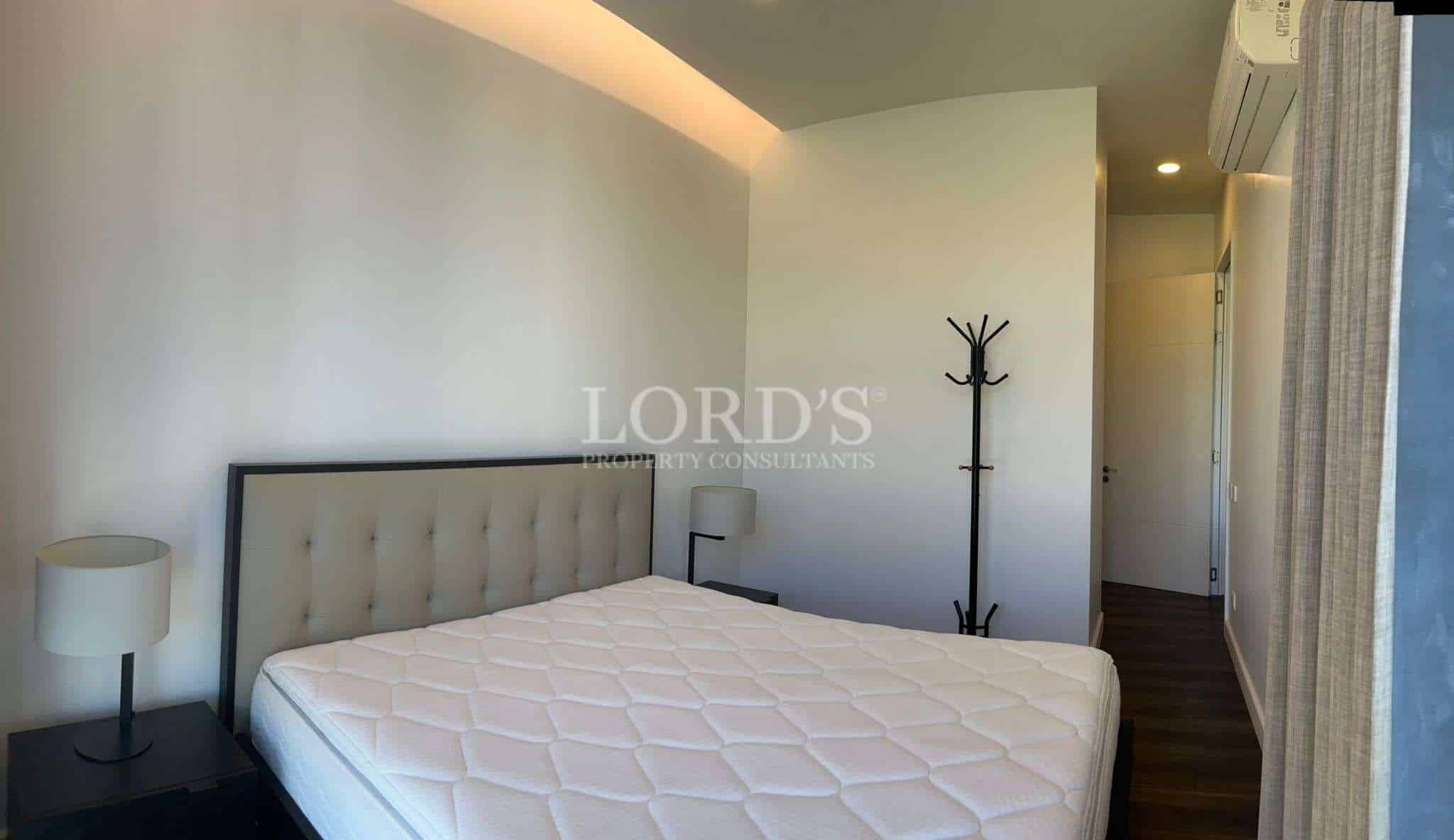 Minimalist bedroom with upholstered headboard, soft lighting, and hallway access in a modern apartment.