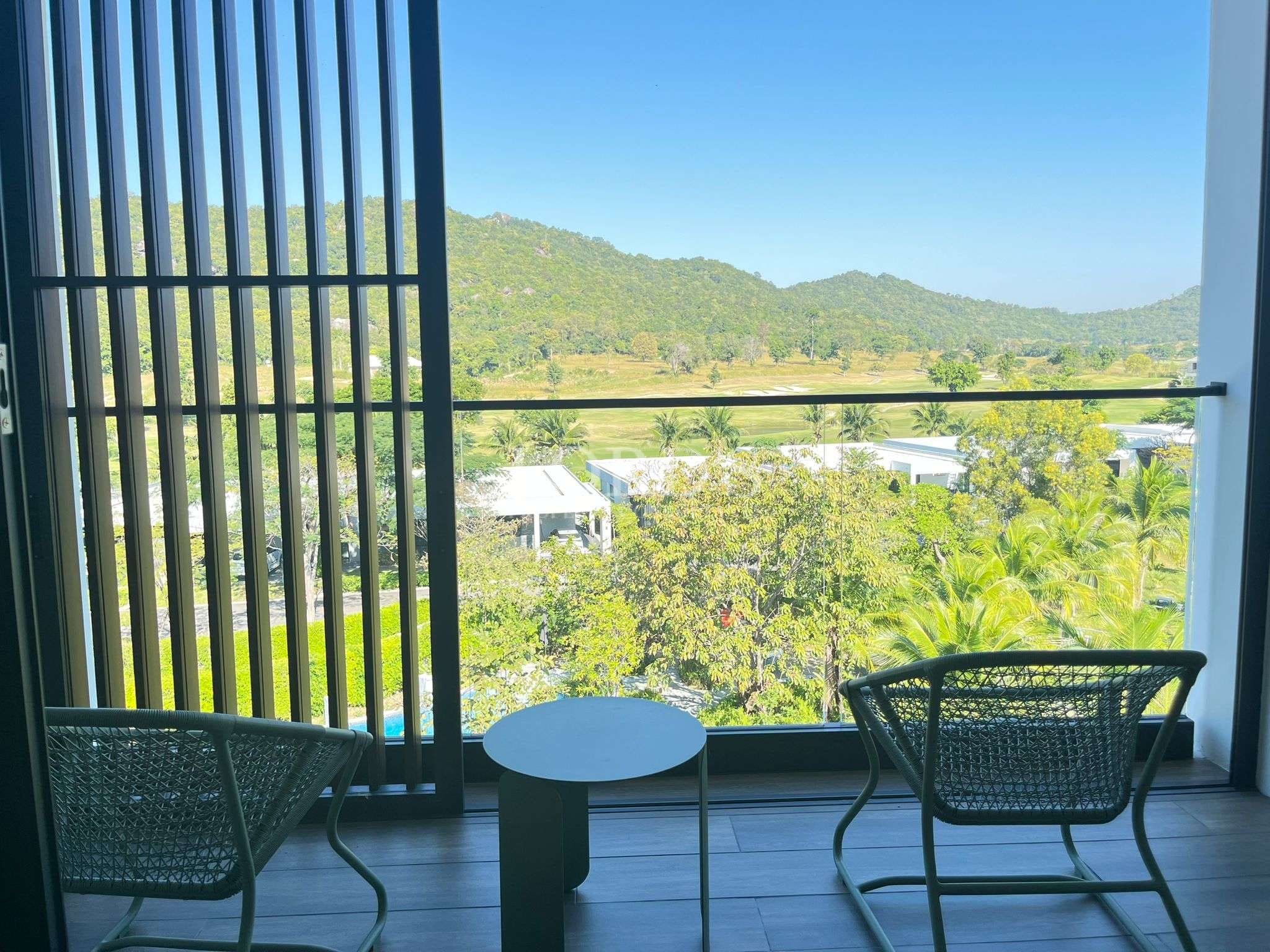 Private balcony with outdoor chairs overlooking lush green hills, palm trees, and a scenic resort-style landscape under a clear blue sky.