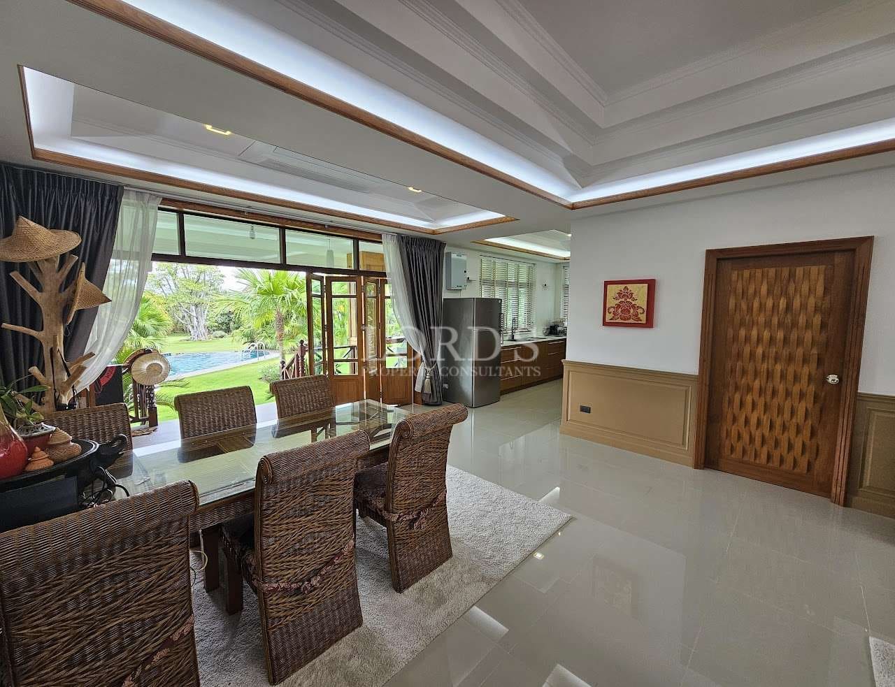 Luxury open-plan dining and living area with wicker dining set, recessed ceiling lighting, wooden door, and garden view through large glass doors