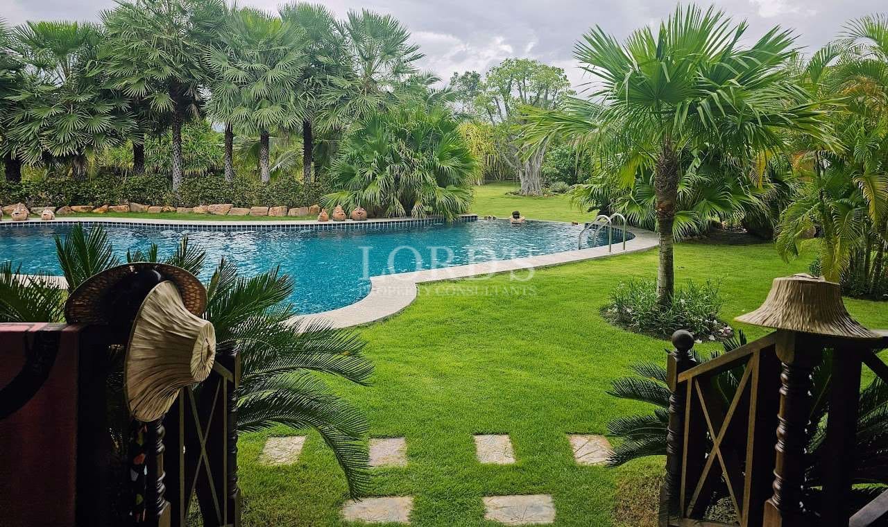 Tropical garden swimming pool with curved edge, palm trees, and manicured green lawn viewed from a terrace.