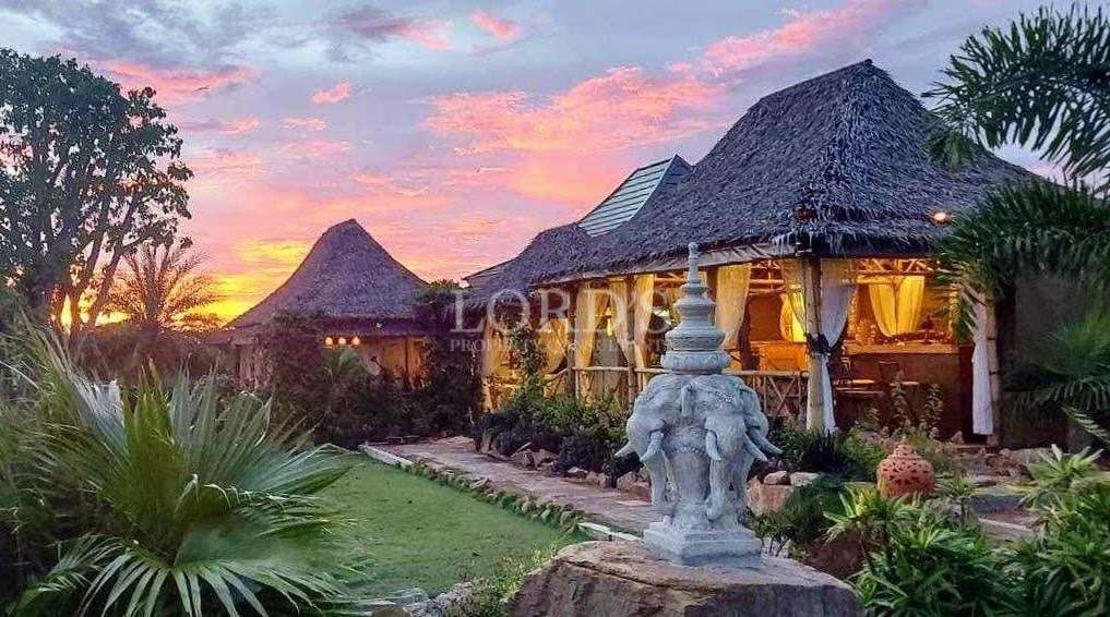 Luxury tropical villa with traditional thatched roof, lush garden landscaping, stone statue, and colorful sunset sky