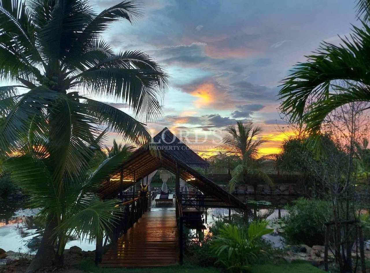 Tropical pavilion over water at sunset, surrounded by palm trees and reflective lagoon.