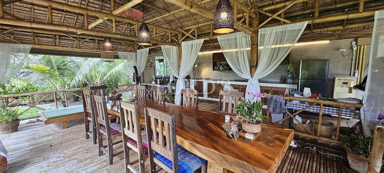 Open-air bamboo dining and kitchen pavilion with a long wooden table, white sheer curtains, pendant lighting, and tropical garden views.
