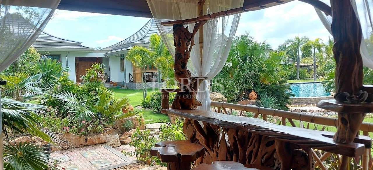 Covered outdoor pavilion with carved wooden seating overlooking a tropical garden and swimming pool at a private villa.