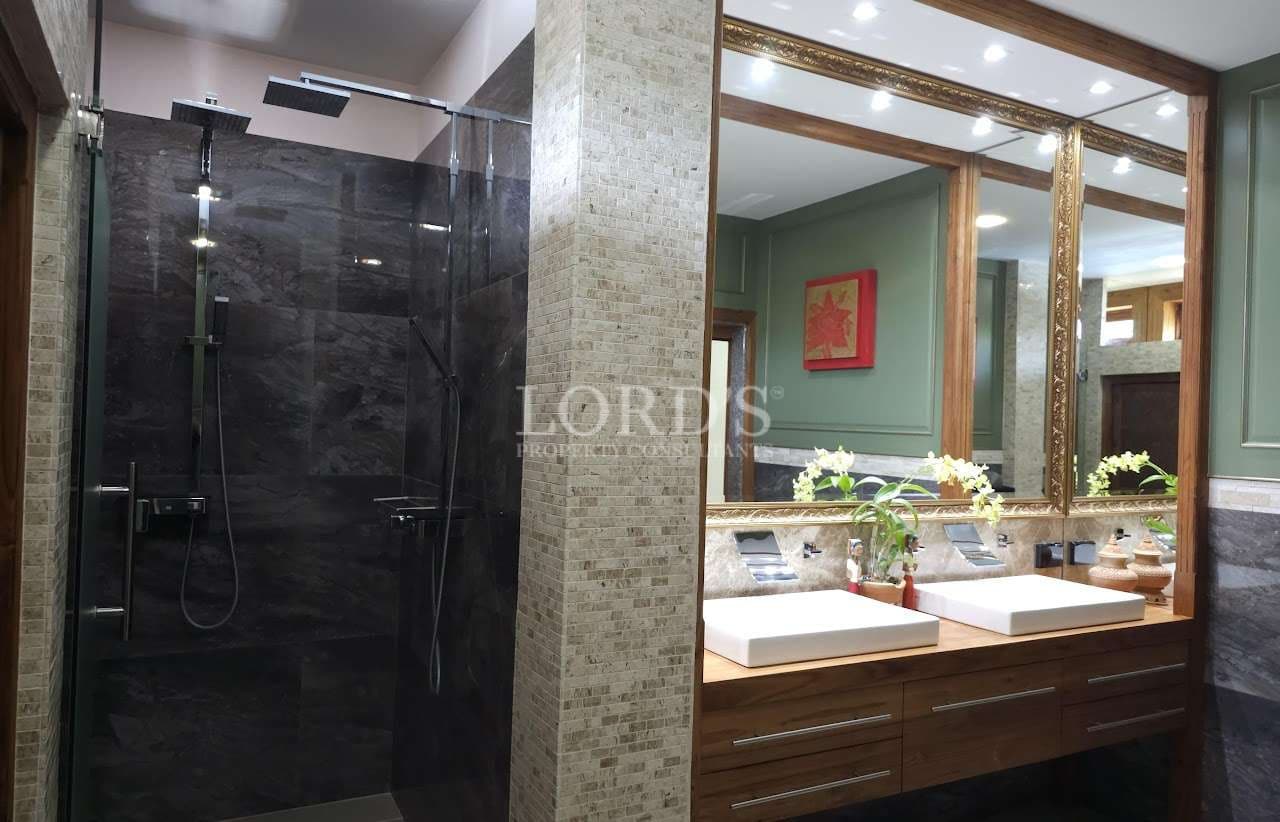 Luxury bathroom with glass-enclosed rainfall shower, double vessel sink vanity, wooden cabinetry, stone walls, and gold-framed mirrors