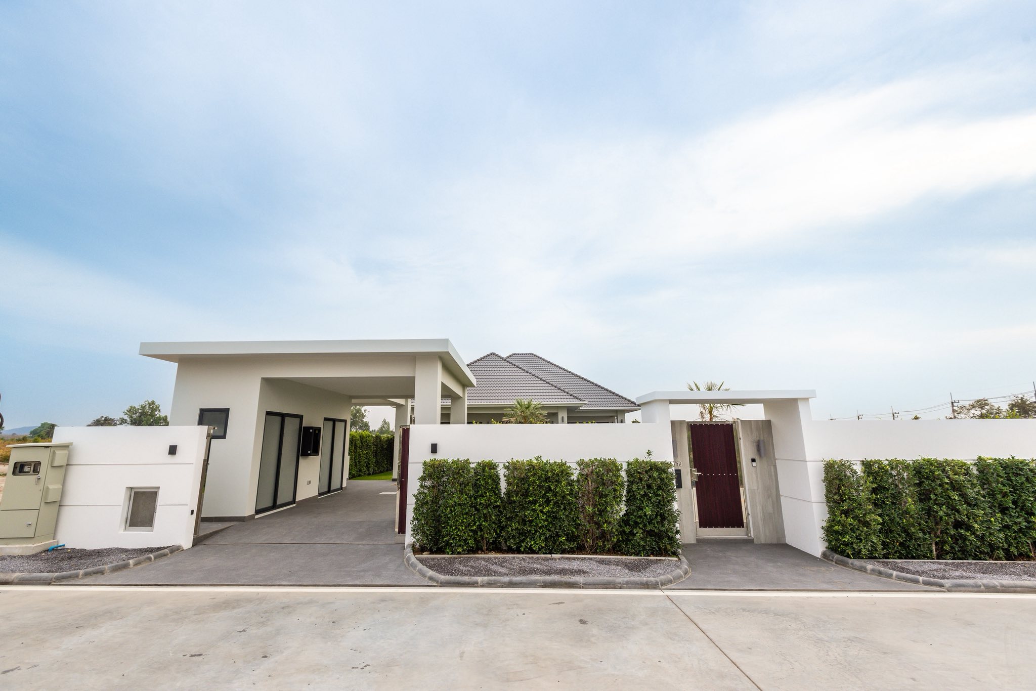 Modern single-story house exterior with white boundary wall, wooden gate, and landscaped hedges.