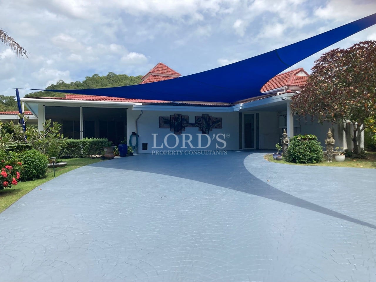Single-storey villa with wide curved driveway, blue shade sail, covered parking, and landscaped tropical garden.