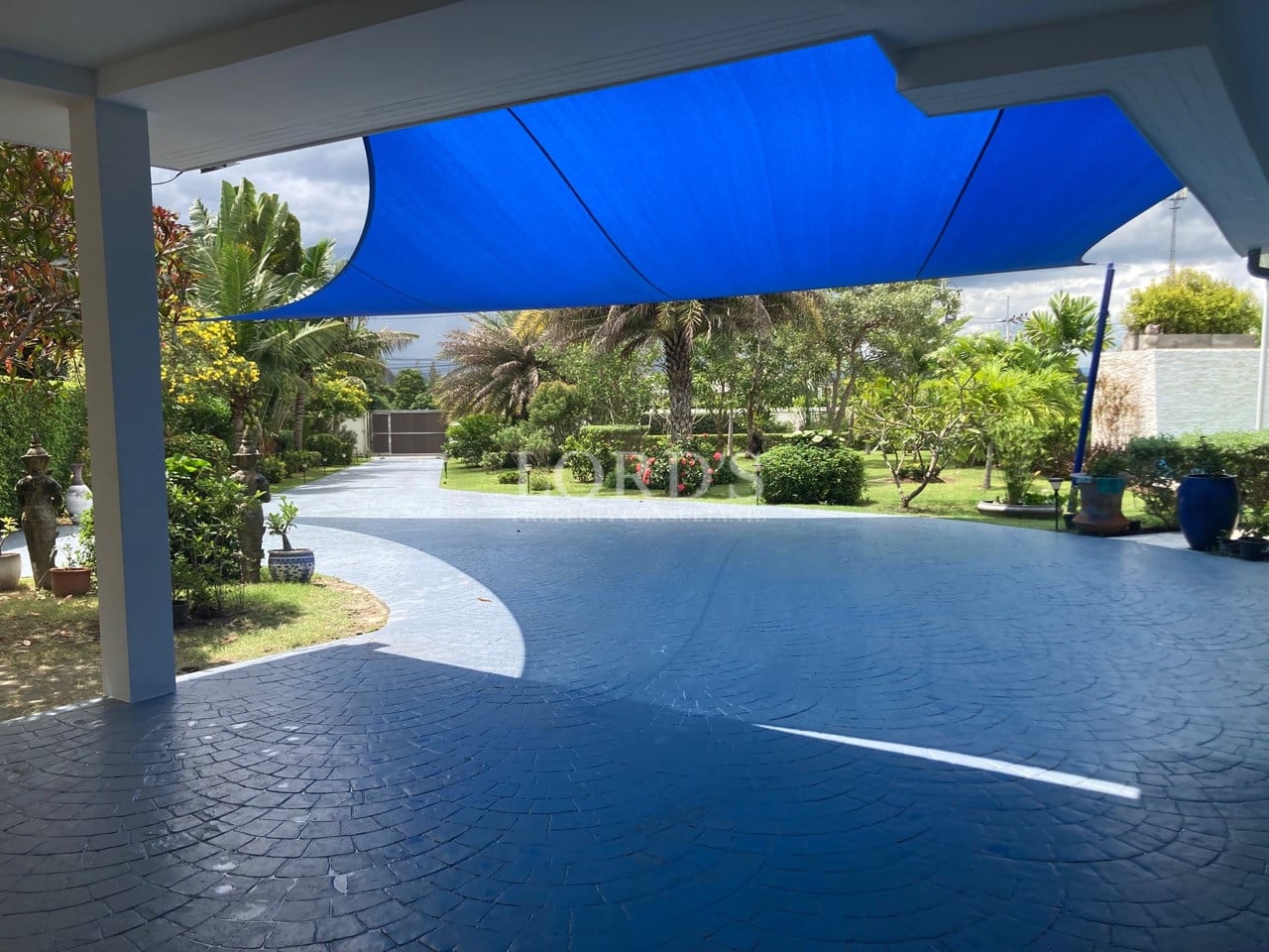 Spacious covered driveway with blue shade sail, decorative paving, and landscaped tropical garden.