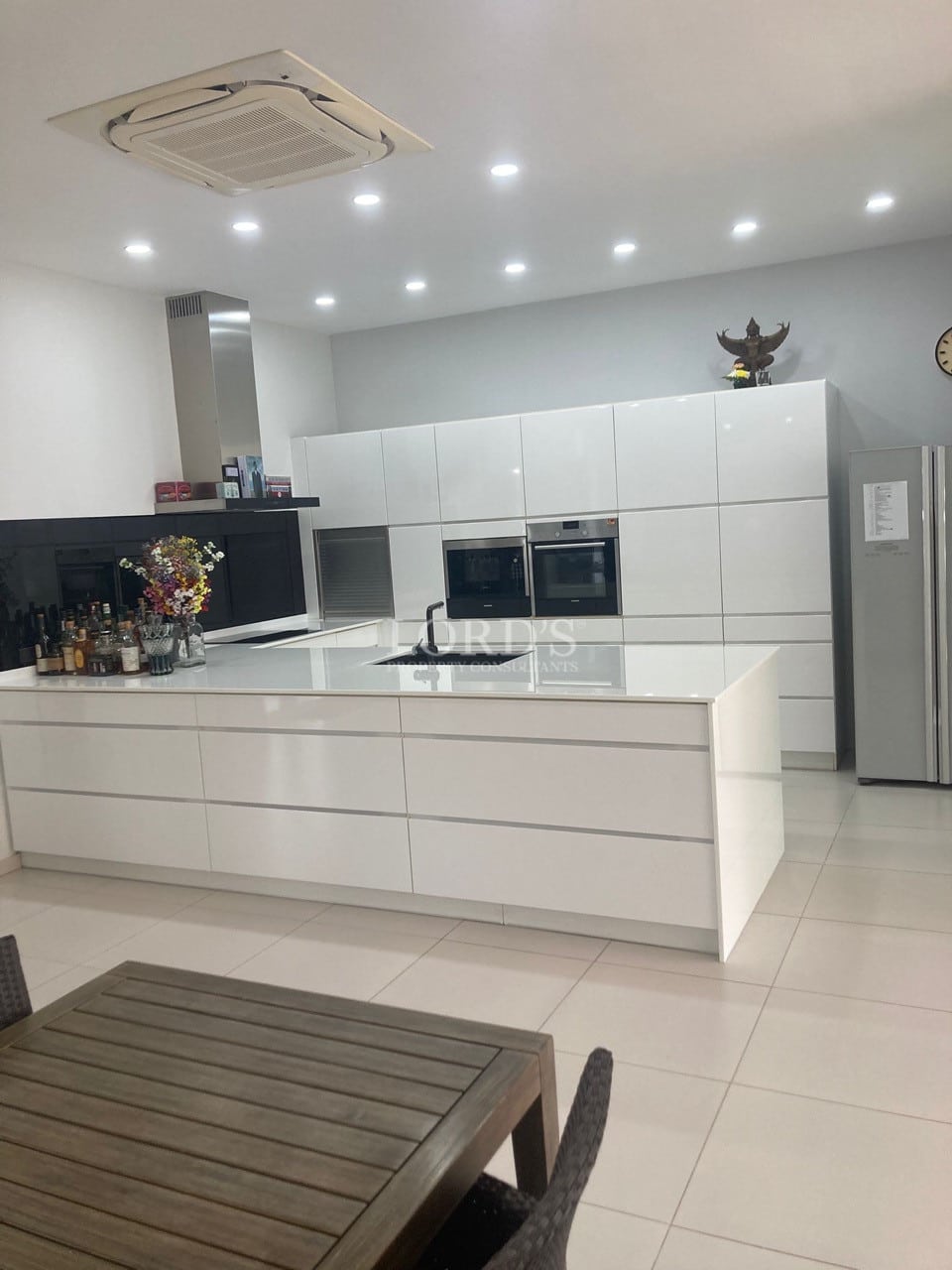 Modern white kitchen with central island, built-in appliances, recessed lighting, and minimalist cabinetry.